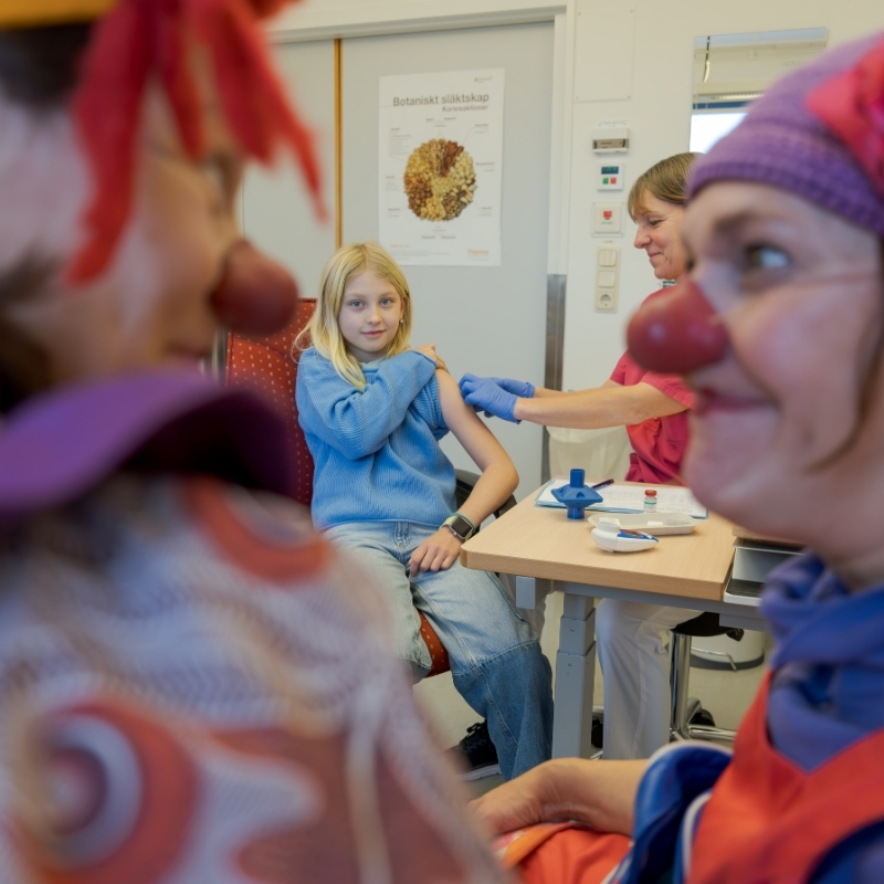 Foto på ett barn som ska ta en spruta på sjukhus. Barnet tittar uppmärksamt på två sjukhusclowner.
