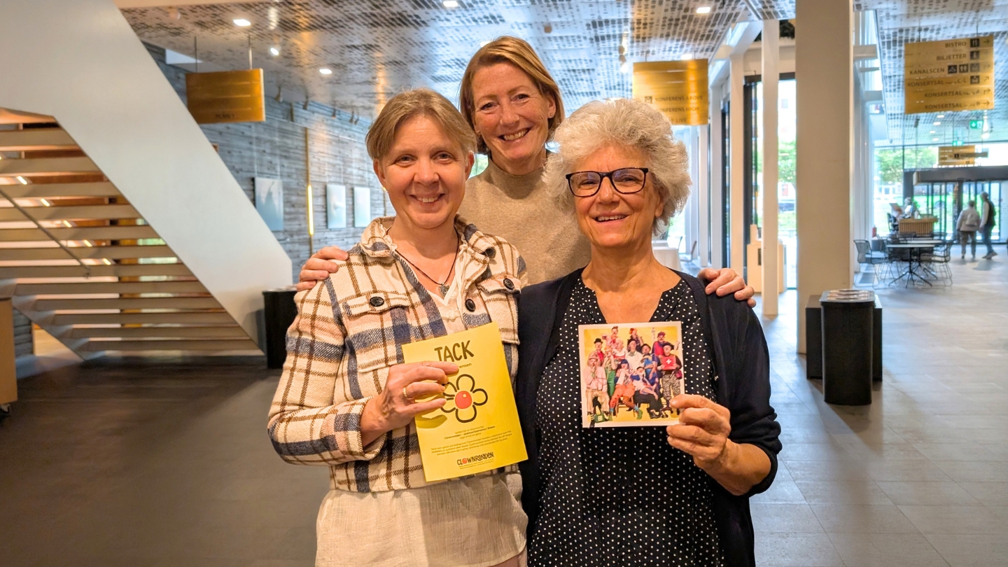 Foto på tre kvinnor som står i en eventlokal. De håller upp ett gåvobevis och ett vykort från Clownronden.