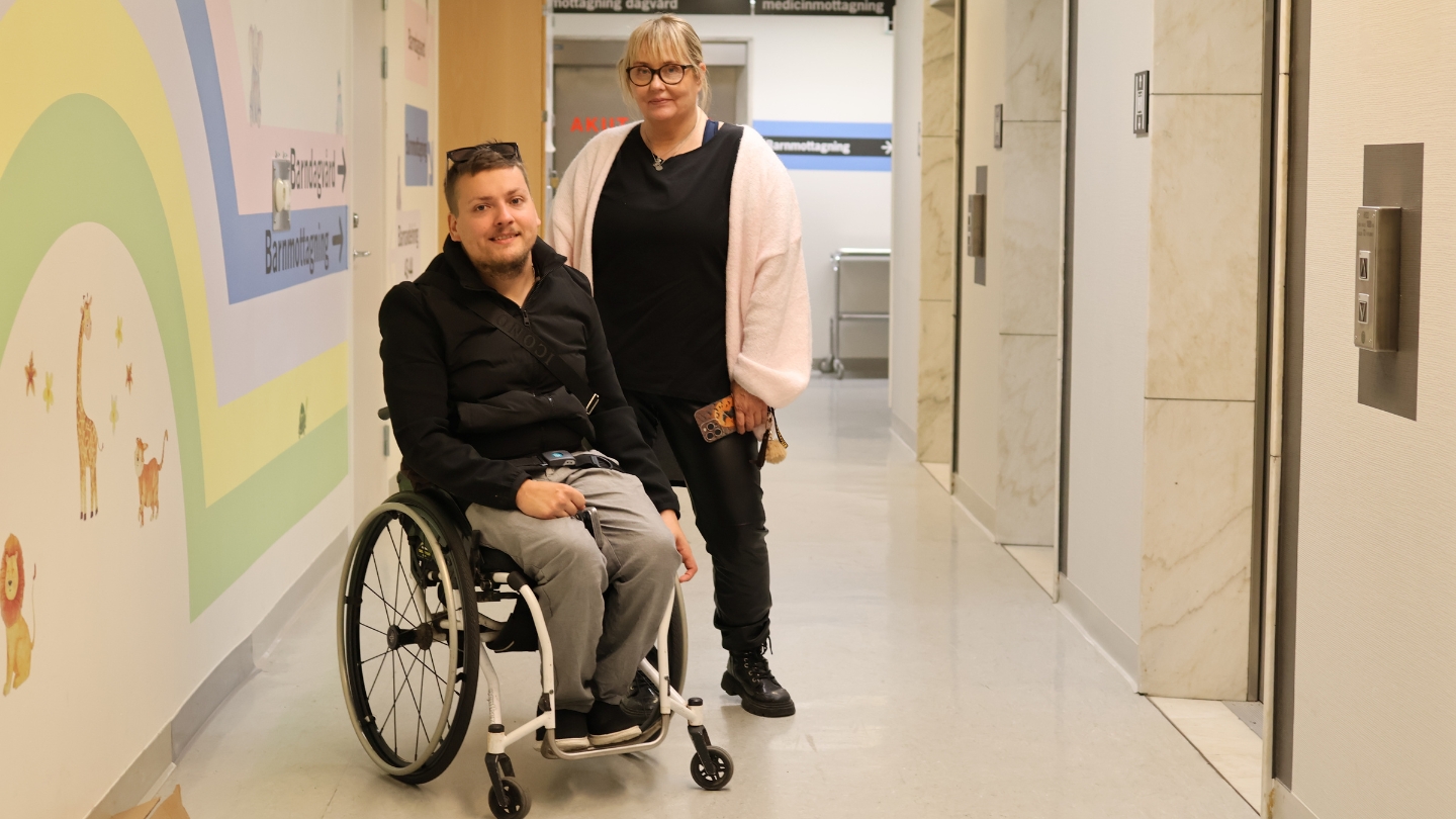 Foto på mor och son i en hisshall på ett sjukhus. Den vuxne sonen sitter i rullstol och mamman står bakom. I bakgrunden syns skyltar till sjukhusets barnavdelningar.